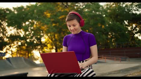 Young-short-haired-girl-in-a-purple-top-finishes-her-job-online-in-the-park.-The-girl-takes-off-the-red-headphones-and-closes-the-red-laptop-in-the-park