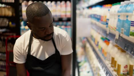 a-Black-man-in-a-white-t-shirt-and-black-apron-laying-out-bottles-of-milk-on-the-counter-of-a-supermarket-with-dairy-products