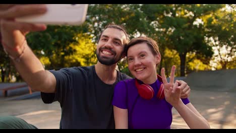 Happy-couple-brunette-guy-in-a-gray-t-shirt-and-a-girl-with-a-short-haircut-in-a-purple-top-with-red-headphones-make-faces-and-take-a-selfie-using-a-white-phone-in-the-park