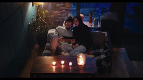 Romantic-evening:-a-brunette-guy-and-a-girl-are-sitting-on-the-couch-and-looking-at-the-screen-of-the-tablet-in-the-evening.-There-are-candles-on-the-table