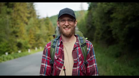 A-blond-man-tourist-with-a-beard-in-glasses-in-a-cap-stands-in-a-plaid-red-shirt-near-the-road-against-the-backdrop-of-the-forest