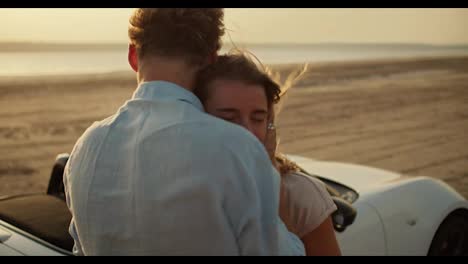 A-happy-girl-is-hugging-her-boyfriend-in-a-blue-shirt-near-a-white-car.-A-romantic-summer-meeting-against-the-river-and-the-yellow-sky