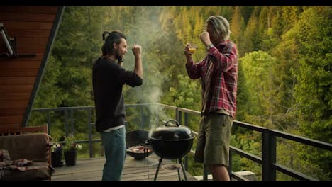 Two-guys-dance-and-have-fun-while-cooking-on-the-grill-on-a-spacious-balcony-overlooking-coniferous-ice-and-mountains.-Picnic-fun,-happy-leisure