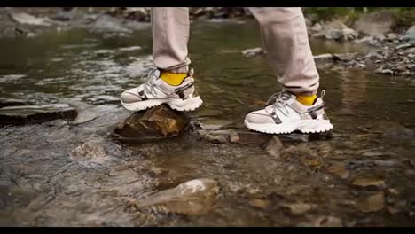 Shooting-close-up:-a-girl-in-white-sneakers-crosses-a-mountain-river-along-a-special-path-made-of-stones,-her-boyfriend-who-is-standing-on-the-other-side-of-the-river-helps-her