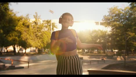 Portrait-of-a-girl-with-a-short-haircut-in-a-purple-top,-striped-pants-and-red-headphones-is-dancing-on-roller-skates-in-a-skatepark-at-sunset-in-summer