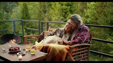 A-blond-man-in-glasses-and-a-blond-girl-are-sitting-on-a-sofa-and-stroking-their-big-light-colored-dog-on-the-balcony-of-a-country-house-overlooking-the-forest-and-mountains