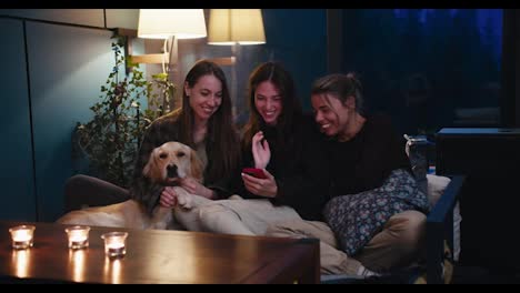 Three-girls-are-sitting-on-the-sofa-and-talking-in-a-cozy-evening-room,-next-to-them-is-their-light-colored-dog.-Candles-on-the-table-and-yellow-lamp-lighting