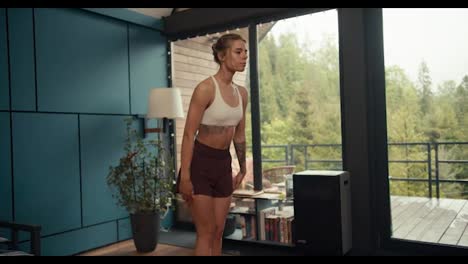 A-blonde-girl-in-a-white-top-with-tattoos-does-balance-and-coordination-exercises-on-a-special-mat-in-an-industrial-house-overlooking-a-coniferous-forest