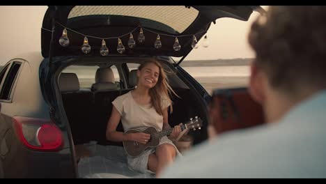 POV:-A-bearded-man-with-curly-hair-in-a-blue-shirt-takes-a-photo-of-his-blonde-girlfriend,-who-is-smiling-and-playing-the-ukulele-in-the-trunk-of-a-black-car-decorated-with-lights-and-shows-her-the-photo-against-a-yellow-sky