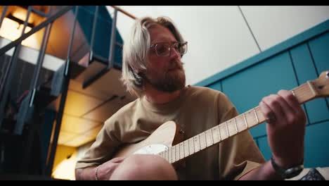 View-from-below:-A-blond-man-with-long-hair-in-glasses-with-a-beard-plays-the-electric-guitar-in-a-cozy-room