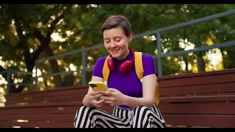 A-girl-with-a-short-haircut-in-a-purple-top,-striped-pants-and-a-yellow-backpack-sits-on-brown-stands-and-chats-on-a-yellow-phone