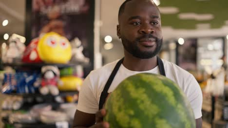 Ein-Schwarzhäutiger-Mann-In-Weißem-T-Shirt-Und-Schwarzer-Schürze-Trägt-Eine-Wassermelone-Und-Stellt-Sie-Auf-Die-Supermarkttheke.-Video-In-Hoher-Qualität-Gefilmt