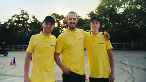 Happy-and-friendly-trio-of-instructors-at-a-driving-school.-Three-guys-in-yellow-T-shirts-posing-in-front-of-motorcycles