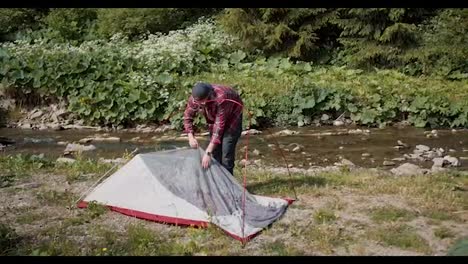 A-review-of-a-guy-in-a-red-plaid-shirt-who-assembles-a-tent-on-his-hike-near-a-mountain-river-in-the-forest