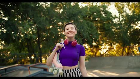 Portrait-of-a-young-short-haired-girl-in-a-purple-top-with-red-headphones-who-poses-and-holds-pink-rollers-in-her-hands-against-the-backdrop-of-a-summer-park