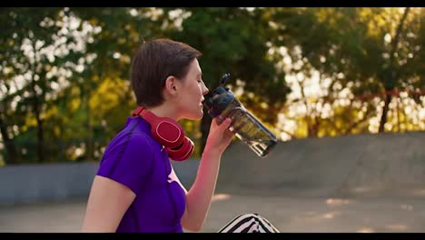 Side-view-of-a-girl-in-a-purple-top-and-red-headphones-in-a-short-haircut-drinks-special-water-in-a-skate-park-in-summer