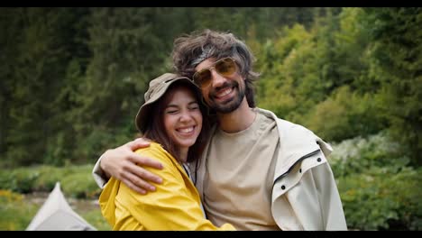 Portrait-of-a-happy-couple:-a-guy-and-a-girl-are-standing-in-hiking-clothes,-hugging-and-smiling-against-the-backdrop-of-a-green-forest