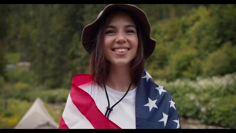 Ein-Glückliches-Brünettes-Mädchen-Mit-Grünem-Hut,-Das-In-Die-Flagge-Der-Vereinigten-Staaten-Von-Amerika-Gehüllt-Ist,-Lächelt-Und-Blickt-Vor-Dem-Hintergrund-Eines-Grünen-Waldes-Und-Eines-Gebirgsflusses-In-Die-Kamera