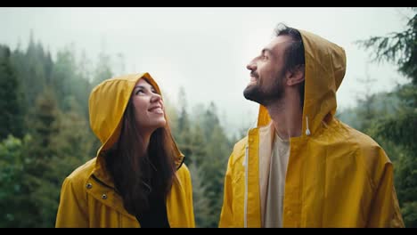 A-happy-couple:-a-guy-and-a-girl-in-yellow-jackets-stand-against-the-backdrop-of-a-foggy-mountain-forest,-smile-and-wait-for-the-coming-rain.-Boy-and-girl-look-at-the-sky-and-hug