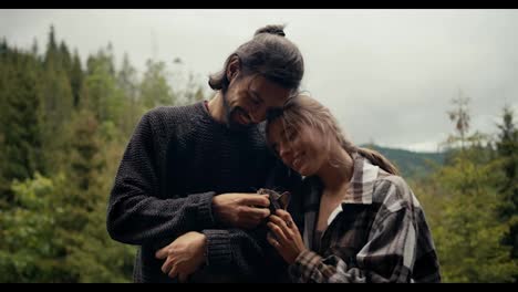 A-guy-in-a-blue-sweater-and-a-blonde-girl-are-stroking-a-cat,-which-is-being-held-by-a-brunette-guy.-Rest-with-a-pet-in-a-country-house-overlooking-a-coniferous-mountain-forest