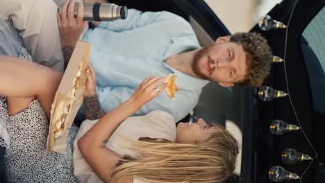 Vertical-video:-A-blonde-girl-gives-her-bearded-boyfriend-with-curly-hair-in-a-blue-shirt-a-taste-of-pizza,-after-which-he