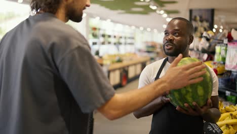 a-man-with-Black-skin-in-a-black-apron-holds-a-watermelon-in-his-hands-and-talks-about-it.-A-store-customer-asks-a-consultant-about-watermelon