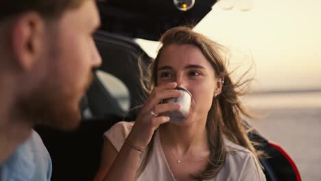 POV:-Blonde-girl-listening-to-her-bearded-boyfriend-while-sitting-in-the-trunk-of-a-black-car,-drinking-tea-and-laughing-against