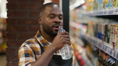 Ein-Mann-Mit-Schwarzer-Hautfarbe-Im-Karierten-Hemd-Trinkt-In-Einem-Supermarkt-Wasser-Aus-Einer-Durchsichtigen-Plastikflasche