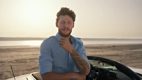 Portrait:-A-bearded-man-with-curly-hair-wearing-a-blue-shirt-is-crosses-his-arms-over-his-chest-and-shows-his-tattoos,-standing-next-to-a-white-convertible-against-the-yellow-sky-on-the-beach