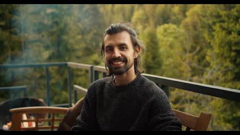A-young-man-in-a-green-sweater-looks-at-the-camera-and-smiles-while-sitting-on-the-sofa-against-the-backdrop-of-mountains-and-coniferous-forest