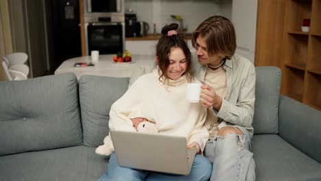 Young-adult-couple-talking-using-laptop-computer-at-home-sitting-on-couch,-watching-video