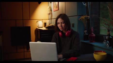 A-happy-brunette-girl-in-red-headphones-rejoices-and-looks-at-the-screen-of-a-white-laptop-in-a-dark-evening-room-illuminated-by-a-yellow-lamp
