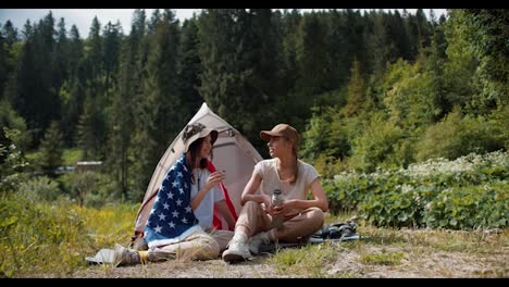 A-blonde-girl-and-a-brunette-girl-in-a-hat-who-swam-in-the-American-flag-sit-near-the-tent-against-the-backdrop-of-a-green-forest-and-communicate