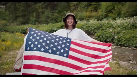 Retrato-De-Una-Chica-Morena-Patriótica-Con-Una-Camiseta-Blanca-Que-Sostiene-La-Bandera-De-Los-Estados-Unidos-De-América-Sonríe-En-El-Contexto-De-Un-Bosque-Verde-Y-Un-Río-De-Montaña