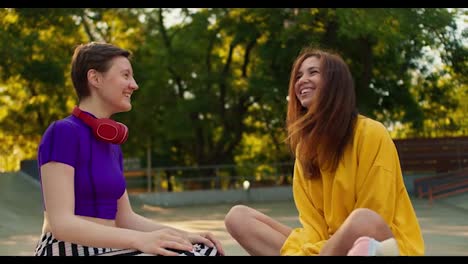 A-brunette-girl-in-a-white-sweater-and-a-girl-with-a-short-haircut-in-a-purple-top-and-red-headphones-communicate-while-sitting-on-the-floor-in-a-skate-park-in-summer