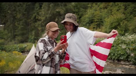 A-blonde-girl-in-a-cap-shows-a-brunette-girl-in-a-white-T-shirt-with-an-American-flag,-a-photo-that-was-taken-during-a-hike-in-the-forest