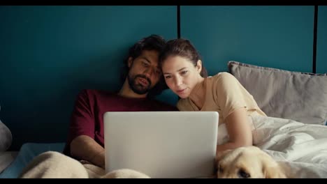 The-guy-and-the-girl-are-looking-at-the-laptop-screen-while-lying-on-the-bed-against-the-background-of-a-turquoise-wall-in-the-morning.-The-couple-jointly-decides-on-the-purchase-of-goods-in-the-online-store