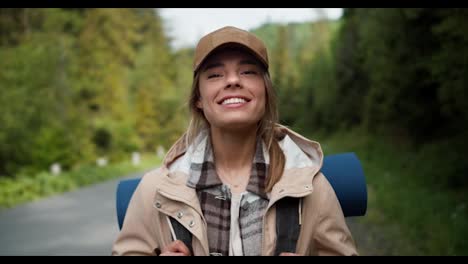 Portrait-of-a-blonde-girl-in-a-cap-in-a-special-uniform-for-hiking-smiling-looking-at-the-camera-against-the-backdrop-of-a-mountain-forest-near-the-road