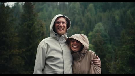 Happy-couple:-a-blond-guy-in-glasses-with-a-beard-and-a-blond-girl-in-jackets-stand-and-rejoice-against-the-backdrop-of-a-green-forest-in-the-mountains