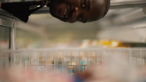 Close-up-shot-of-a-Black-skinned-man-in-a-plaid-shirt-looks-into-the-supermarket-refrigerator,-looks-into-the-camera-and-takes-an-ice-cream