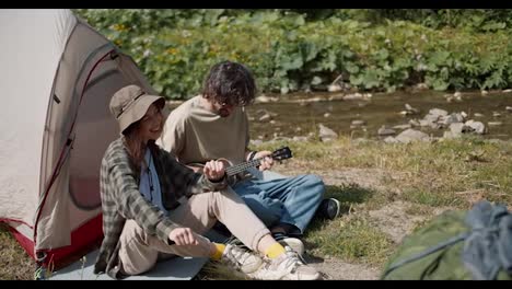 Shooting-near-a-man-and-a-girl-are-sitting-near-a-tent-a-man-is-playing-a-guitar-A-girl-is-listening-to-him-and-dancing-they-are-sitting-near-a-mountain-river