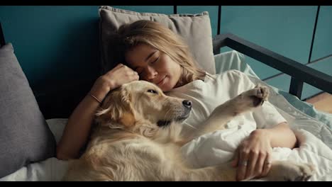 Close-up-shot:-a-blonde-girl-sleeps-in-bed-with-her-light-colored-dog-in-the-morning.-Happy-pet-owner