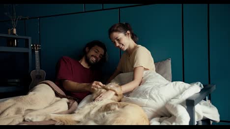 A-brunette-guy-in-a-red-T-shirt-and-a-brunette-girl-communicate-and-stroke-their-dog-in-bed-against-the-background-of-a-turquoise-wall-at-home