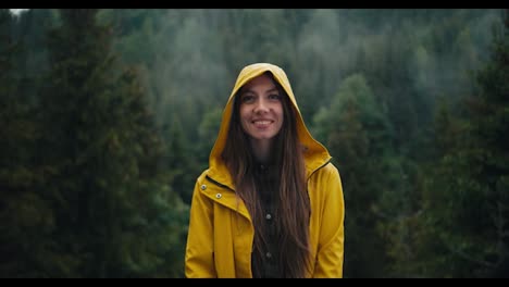 A-happy-girl-in-a-yellow-jacket-stands-and-smiles-against-the-backdrop-of-a-green-coniferous-forest-in-the-mountains-during-the-rain