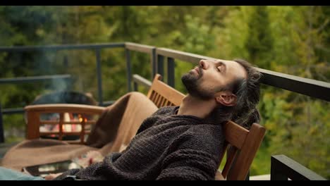 A-young-brunette-man-with-closed-eyes-is-resting-on-a-sofa-on-the-balcony-of-a-country-house-overlooking-the-mountains-and-a-coniferous-forest,-firewood-is-burning-near-him-in-the-barbecue