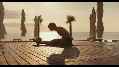 The-guy-does-yoga-exercises-on-a-sunny-beach-in-the-morning,-flooring-with-boards.-Morning-exercise-on-the-beach-at-sunrise