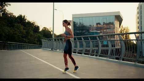 A-sports-girl-in-a-sports-summer-uniform-runs-in-place-and-warms-up-before-a-morning-run-against-the-background-of-a-bridge-in-the-city