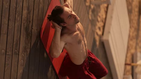 Vertical-video:-a-brunette-guy-with-a-naked-torso-in-red-pants-gives-himself-a-back-massage-with-a-cone-and-relaxes-after-a-yoga-class-on-a-red-mat-on-a-sunny-beach-in-the-summer
