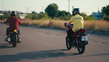 An-instructor-in-a-yellow-T-shirt-and-helmet-rides-Near-his-student-in-a-red-T-shirt-and-helmet-on-a-motorcycle.-School-day-at-the-driving-school