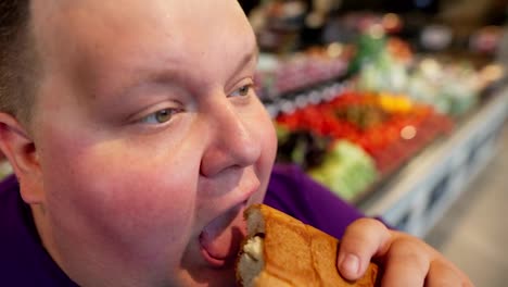 Close-up-shot:-happy-overweight-man-in-purple-t-shirt-eating-croissant-in-supermarket.-Face-of-an-overweight-man-eating-sweets-in-a-supermarket
